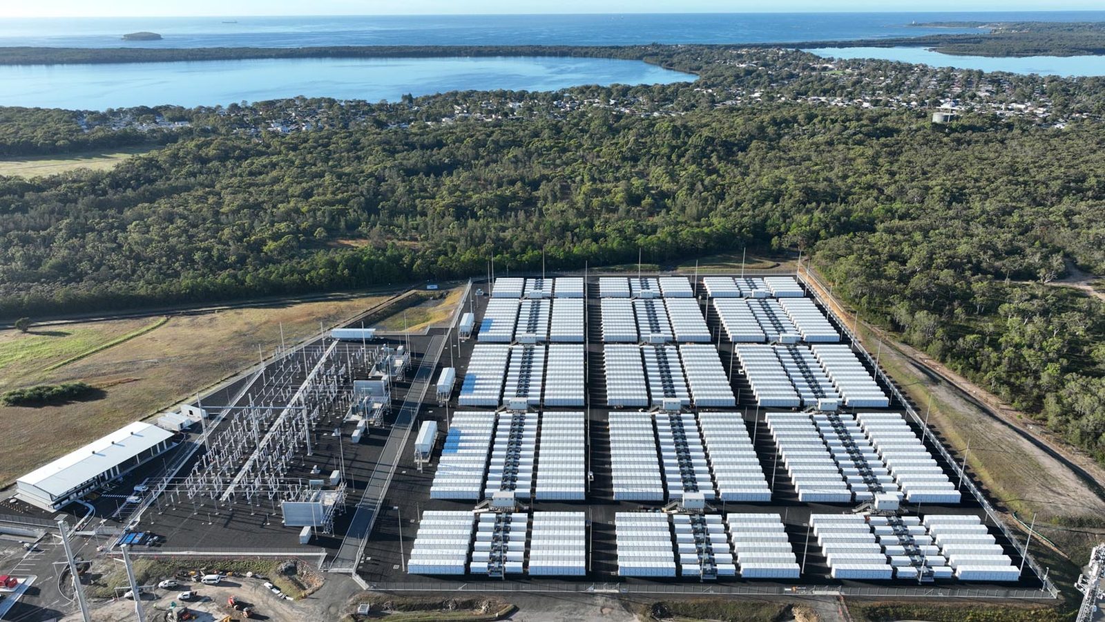 Aerial view of a large grid scale battery storage facility, long rows of white battery containers next to substation infrastructure.